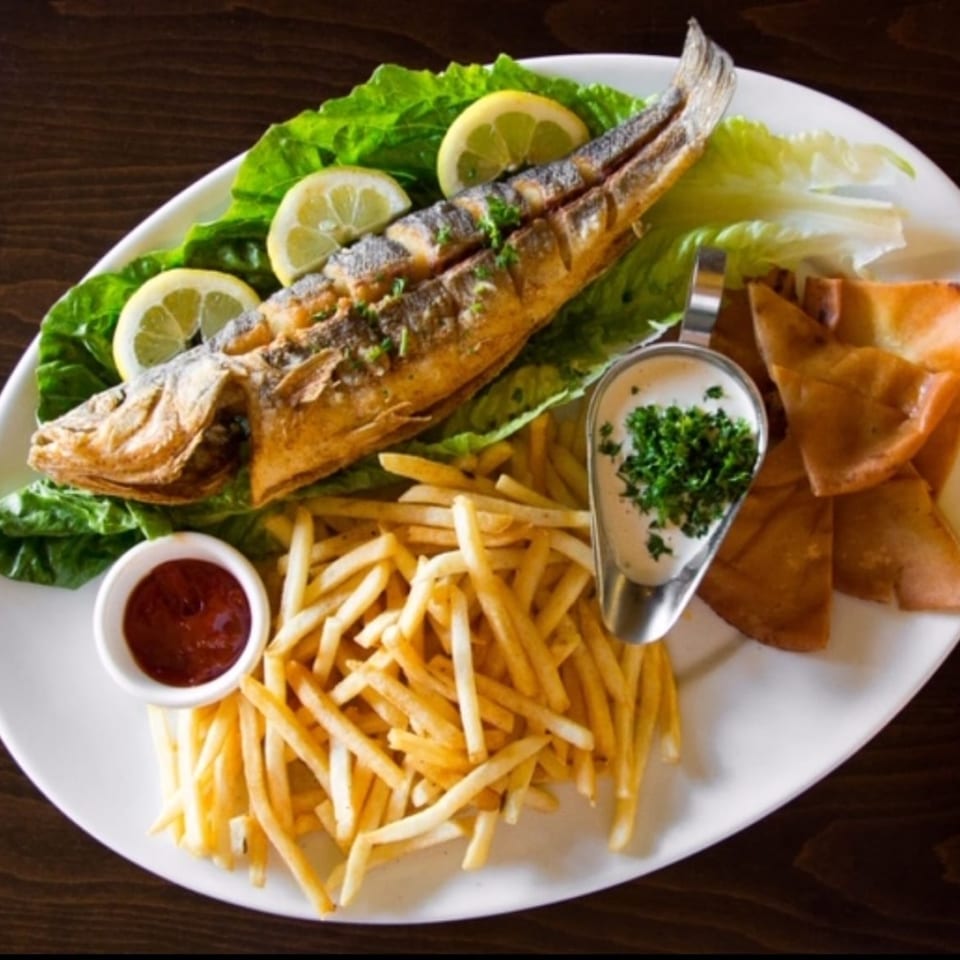 Whole fried Mediterranean fish with lemon, lettuce, fries, ketchup, and pita bread on a white plate.