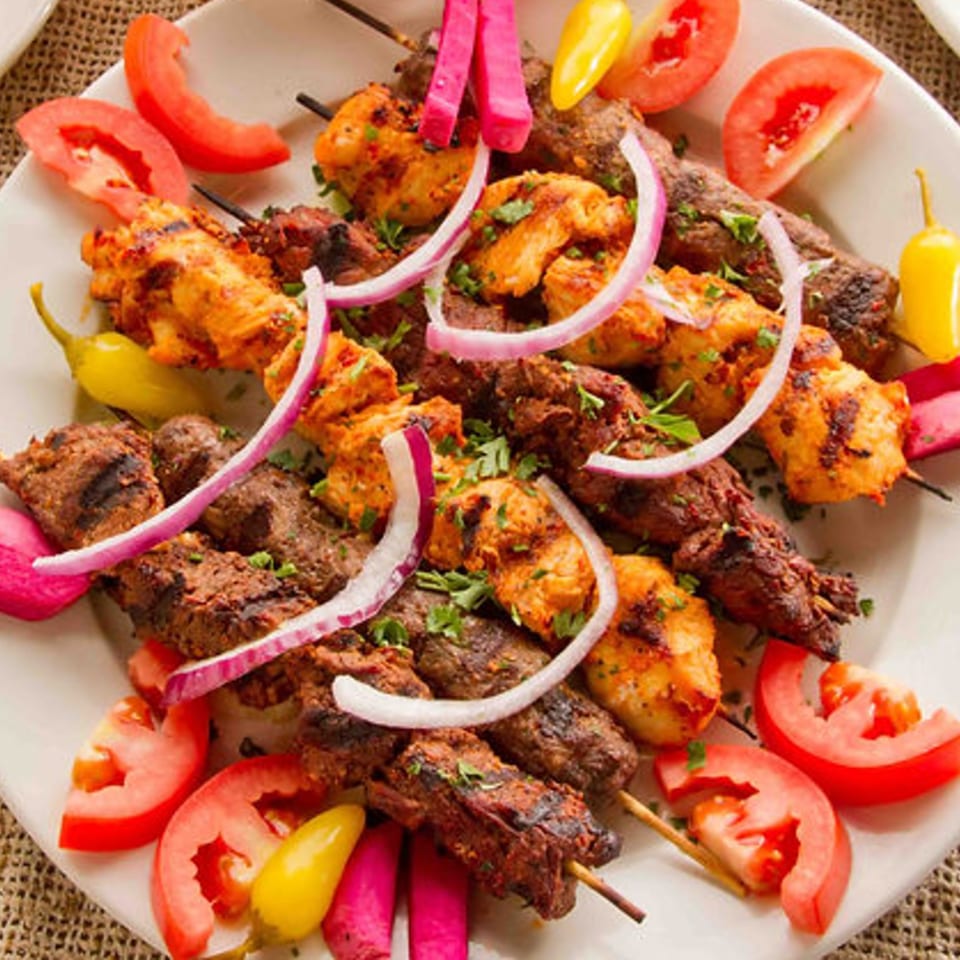 A platter of Mediterranean chicken and beef kebabs with tomatoes, pickled turnips, and onions.