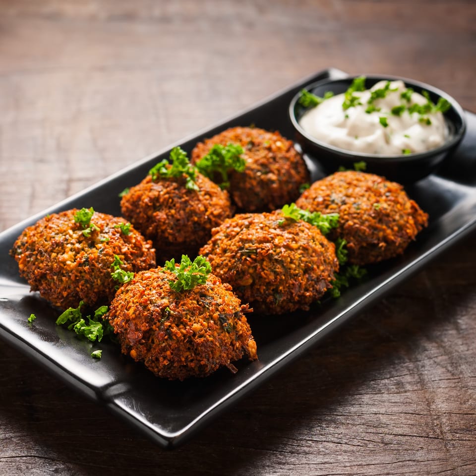 Six falafel with parsley garnish and a side of white dipping sauce on a black plate.