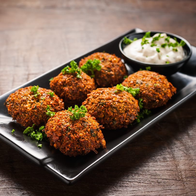 Six falafel with parsley garnish and a side of white dipping sauce on a black plate.